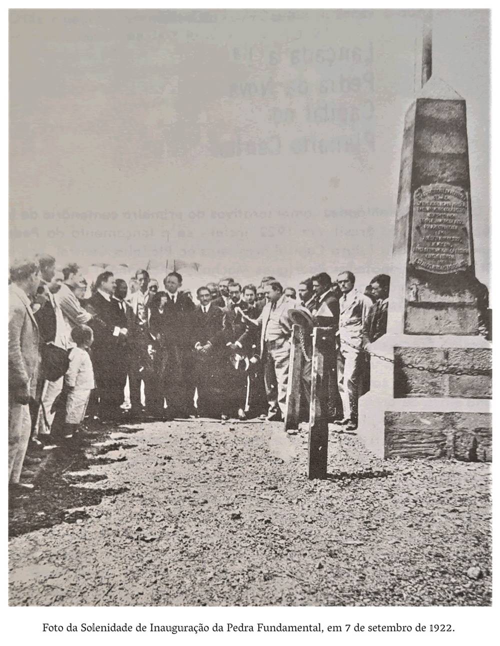  2d- Homens, mulheres e crianças estão reunidos para inauguração da pedra fundamental. Um homem de terno claro, cabelos curtos e bigodes, segura em sua mão direita um papel. Do lado esquerdo um menino vestindo roupas claras e sapatos pretos, olha fixamente para o orador. Realização: Academia Planaltinense de Letras, Artes e Ciências (APLAC), pelo Ecomuseu Pedra Fundamental e pelo Coletivo Nativo Audiodescrição produzida